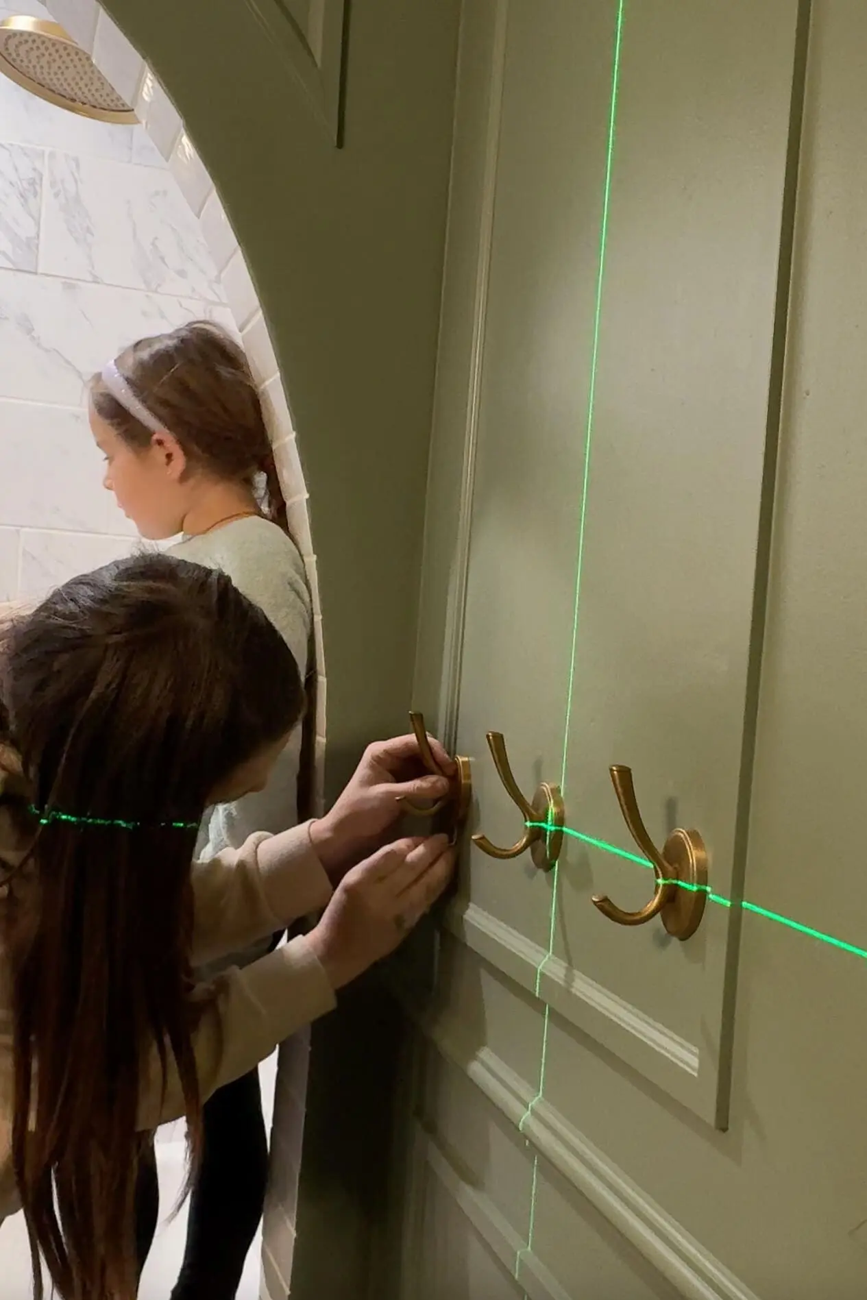 Woman and young girl working together to install brass coat hooks on a dark green bathroom wall, using a laser level to ensure alignment, with a tiled archway and shower in the background.